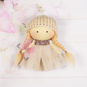 Puede incluir: Una muñeca hecha a mano con trenzas rubias, que lleva un gorro de punto beige, un vestido de tul beige y un cinturón de cuentas marrones. La muñeca sostiene una flor rosa.