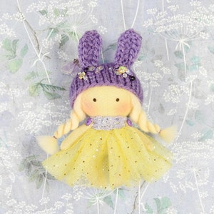 Puede incluir: Una muñeca de tela con cabello de lana rubia en dos trenzas, que lleva un gorro de punto morado con orejas de conejo y pequeños adornos florales. La muñeca tiene una falda de tul amarilla con detalles de purpurina dorada y un cuello de encaje.