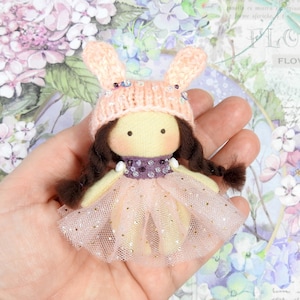 Puede incluir: Una muñeca hecha a mano con un gorro de conejito de punto melocotón claro, trenzas marrones y una falda de tul rosa claro. La muñeca tiene un cuerpo amarillo y un cuello con lentejuelas moradas. La muñeca se sostiene en una mano.