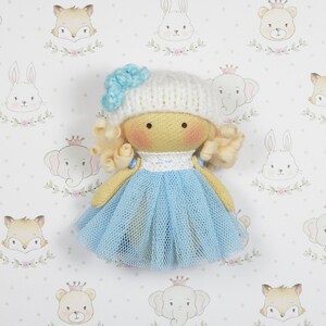 Puede incluir: Una muñeca hecha a mano con cabello rubio, un gorro de punto blanco con una flor azul y un vestido de tul azul claro. La muñeca está sentada sobre un fondo blanco con un patrón repetido de caras de animales.