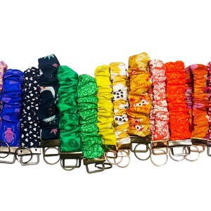 May include: A selection of fabric wrist keychains in a variety of colours and patterns. The keychains display floral, polka dot, and geometric designs. Each keychain has a metal clasp for keys. The colours include pink, purple, blue, green, yellow, and orange.