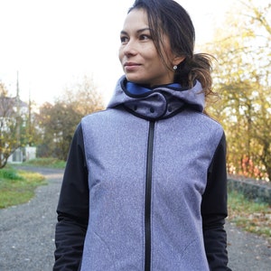 May include: A woman wearing a grey and black hooded jacket with a zip closure. The jacket has a blue lining around the hood and a black zip pull. The jacket has two pockets on the front.