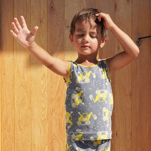 May include: A young child wearing a sleeveless gray tank top with a yellow construction vehicle print. The tank top has a yellow trim around the neckline.