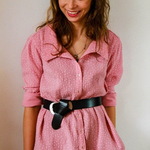 May include: A pink button-down shirt with white polka dots and a black belt with a large silver buckle. The shirt has a wide collar and long sleeves.