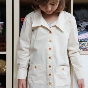 May include: A white button-down shirt with a large collar and pockets. The shirt has gold buttons and is made of a lightweight fabric.
