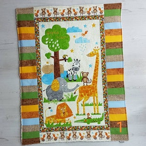 Könnte beinhalten: Eine personalisierte Babydecke mit Dschungel-Motiv. Die Decke zeigt einen grünen Baum, eine Giraffe, einen Elefanten, ein Zebra, einen Löwen, ein Krokodil und Affen. Die Decke ist weiß mit einem braunen, grünen, gelben und blauen Rand. Der Text "Greta 8/10/2014" ist auf die Decke gestickt.