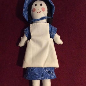 May include: A handmade cloth doll wearing a blue and white dress with a white apron and a blue bonnet. The doll has black hair and a painted face with rosy cheeks.
