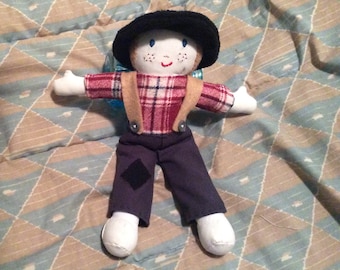 May include: Handmade doll with a black hat, red and black plaid shirt, and blue pants. The doll has white arms and feet, with a black patch on the left knee. It has a smiling face with freckles and is wearing tan suspenders.
