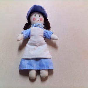 May include: A handmade doll with a blue bonnet, dress, and sleeves, and a white apron. The doll has brown hair, rosy cheeks, and a smiling expression. The doll is made of fabric and has a soft, plush appearance.