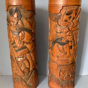 May include: Two tall, intricately carved wooden cylinders with a brown finish. Each cylinder features a detailed relief carving of a samurai warrior on horseback, with intricate details of armour and weaponry.