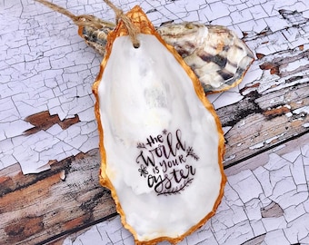 May include: A white oyster shell with gold paint around the edges. The shell has the words "The world is your oyster" written in black calligraphy.