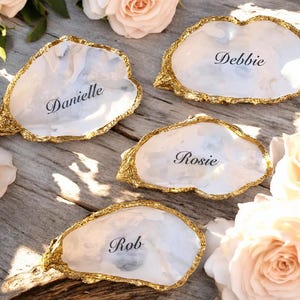 May include: Four white, irregular, shell-shaped trinket dishes with gold-painted edges. Each dish features a black script name printed in the centre: Danielle, Debbie, Rosie, and Rob. The surfaces have a smooth, glossy, marbled appearance.