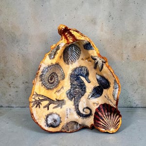 May include: A decorative, seashell-shaped dish with a beige interior featuring illustrations of various sea creatures, including a seahorse, starfish, and shells. The dish has a gold-colored rim and a smaller, red-brown shell attached to the side.