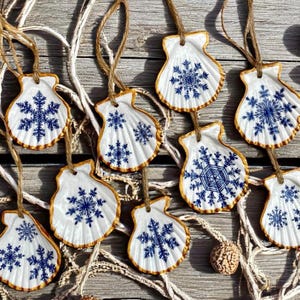May include: A collection of white scallop shell ornaments, each featuring a unique blue snowflake design and a gold-painted edge. Every shell is suspended by a natural jute twine loop, creating a set of hanging decorations with intricate patterns.