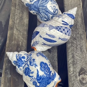 Puede incluir: Tres conchas pintadas con diseños azules y blancos. Una concha presenta un pulpo, otra tiene un patrón de peces y la tercera tiene un diseño de pavo real.