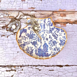 Puede incluir: Un plato de porcelana blanca en forma de concha de mar con un patrón floral azul y blanco. El plato tiene un borde dorado y está decorado con un diseño de pavo real. Un collar de plata con un colgante descansa sobre el plato.