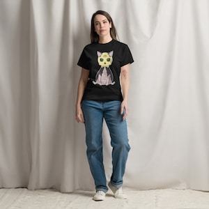 Può includere: Una donna indossa una maglietta nera con una grafica di un gatto rosa con una testa di teschio gialla e occhi verdi. Indossa jeans blu e scarpe da ginnastica bianche.