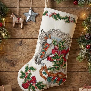 Pode incluir: Uma meia de Natal cor de creme com um design detalhado em ponto cruz de uma cabeça de cavalo adornada com uma rédea vermelha, folhas de azevinho verdes e bagas vermelhas. A meia tem uma borda decorativa de azevinho e bagas no topo.