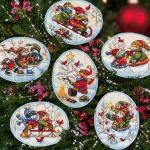 Könnte beinhalten: Sechs ovale Kreuzstich-Ornamente mit verschiedenen Winterszenen, Schneemännern, Vögeln und festlichen Motiven. Jedes Stück ist mit einer hellblauen gedrehten Kordel eingefasst und zeigt farbenfrohe, detaillierte Illustrationen von Schneemännern bei saisonalen Aktivitäten wie Schlittenfahren, Schlittschuhlaufen und dem Schmücken eines Baumes.
