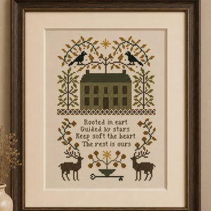 May include: Framed cross-stitch artwork featuring a house, deer, and birds, with the text "Rooted in eart, Guided by stars, Keep soft the heart, The rest is ours." The frame is dark brown.