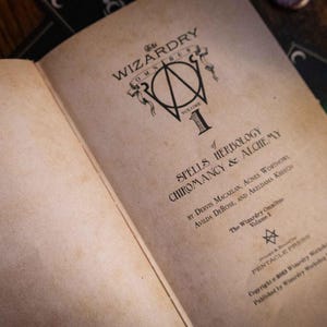 K&ouml;nnte beinhalten: Ein aufgeschlagenes Buch mit gealterten, cremefarbenen Seiten und schwarzem Textdruck. Der Titel lautet The Wizardry Omnibus Volume 1 of Spells Herbology Chiromancy & Alchemy von Dervis Macallan, Agnes Worthorn, Avilda DeRose und Akeldama Kretsch.