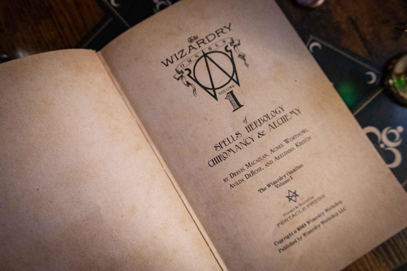 K&ouml;nnte beinhalten: Ein aufgeschlagenes Buch mit gealterten, cremefarbenen Seiten und schwarzem Textdruck. Der Titel lautet The Wizardry Omnibus Volume 1 of Spells Herbology Chiromancy & Alchemy von Dervis Macallan, Agnes Worthorn, Avilda DeRose und Akeldama Kretsch.