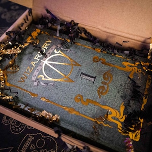 K&ouml;nnte beinhalten: Ein rechteckiges Buch mit dem Titel Wizardry Omnibus Volume 1 mit einem blaugr&uuml;nen Einband, der mit filigranen Goldfolienverzierungen und Schriftz&uuml;gen versehen ist. Das Buch liegt in einem Karton, umgeben von schwarzem und goldenem F&uuml;llmaterial.