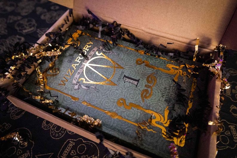 K&ouml;nnte beinhalten: Ein rechteckiges Buch mit dem Titel Wizardry Omnibus Volume 1 mit einem blaugr&uuml;nen Einband, der mit filigranen Goldfolienverzierungen und Schriftz&uuml;gen versehen ist. Das Buch liegt in einem Karton, umgeben von schwarzem und goldenem F&uuml;llmaterial.