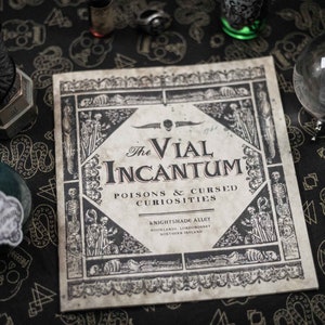 Puede incluir: Un libro de estilo vintage titulado "The Vial Incantum" con el subtítulo "Poisons & Cursed Curiosities." La imagen también presenta una botella de vidrio, una bola de cristal y un adorno de cuervo. El fondo es una tela negra con un patrón de calaveras y serpientes.