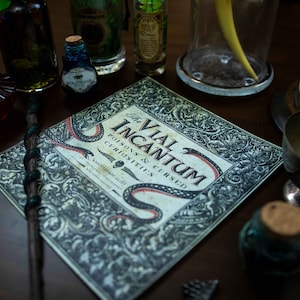 Op de afbeelding: Een boek getiteld "The Vial Incantum: Poisons & Cursed Curiosities" met een zwart slangontwerp op de omslag. Het boek is omringd door verschillende glazen flessen en potten, waaronder een schedel in een pot.