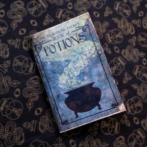 May include: A rectangular book titled The Practical Pocket Book of Potions with a blue and green distressed cover featuring a black silhouette of a cauldron and various mystical symbols rising from it in a cloud-like pattern.
