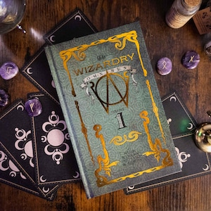 K&ouml;nnte beinhalten: Ein gebundenes Buch mit dem Titel The Wizardry Omnibus Volume 1, einem strukturierten gr&uuml;nen Einband sowie goldener Schrift und dekorativen Verzierungen. Das Buch ist von mehreren schwarzen rechteckigen Karten mit wei&szlig;en Mondsymbolen umgeben.