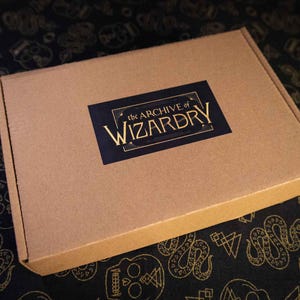 K&ouml;nnte beinhalten: Ein rechteckiger Versandkarton aus brauner Pappe mit einem schwarzen, rechteckigen Aufkleber in der Mitte des Deckels. Das Etikett zeigt den Text The Archive of Wizardry in einer goldenen Serifenschrift mit dekorativen goldenen Linien.