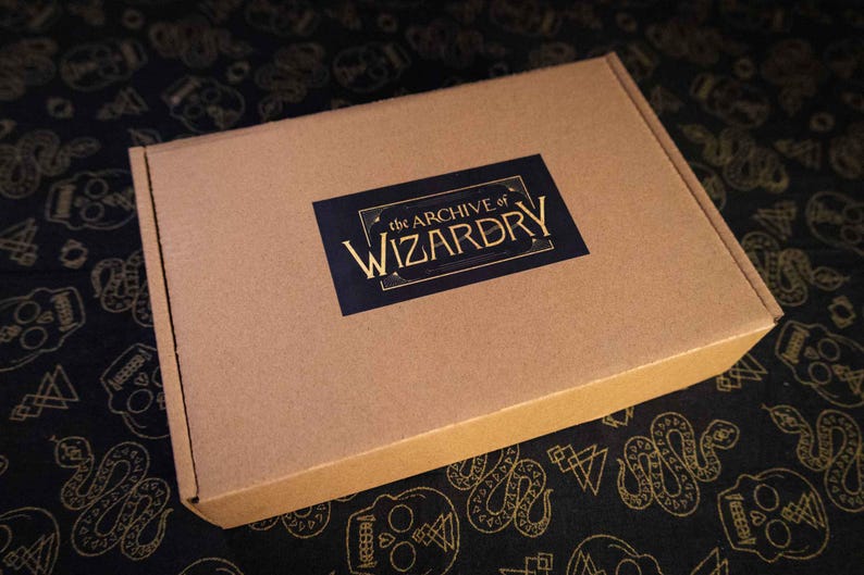 K&ouml;nnte beinhalten: Ein rechteckiger Versandkarton aus brauner Pappe mit einem schwarzen, rechteckigen Aufkleber in der Mitte des Deckels. Das Etikett zeigt den Text The Archive of Wizardry in einer goldenen Serifenschrift mit dekorativen goldenen Linien.