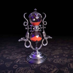 May include: A silver-toned metal hourglass featuring two dragon figures with coiled tails and wings flanking the glass bulbs. The hourglass contains bright orange sand and is supported by a circular base, with intricate dragon head details on each side.