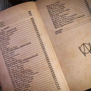 K&ouml;nnte beinhalten: Ein aufgeschlagenes Buch mit gealterten, beigefarbenen Seiten, das ein Inhaltsverzeichnis f&uuml;r Teil IV Alchemie zeigt. Der Text listet verschiedene alchemistische Themen und Seitenzahlen auf. Ein schwarzes Kreissymbol ist rechts abgebildet.
