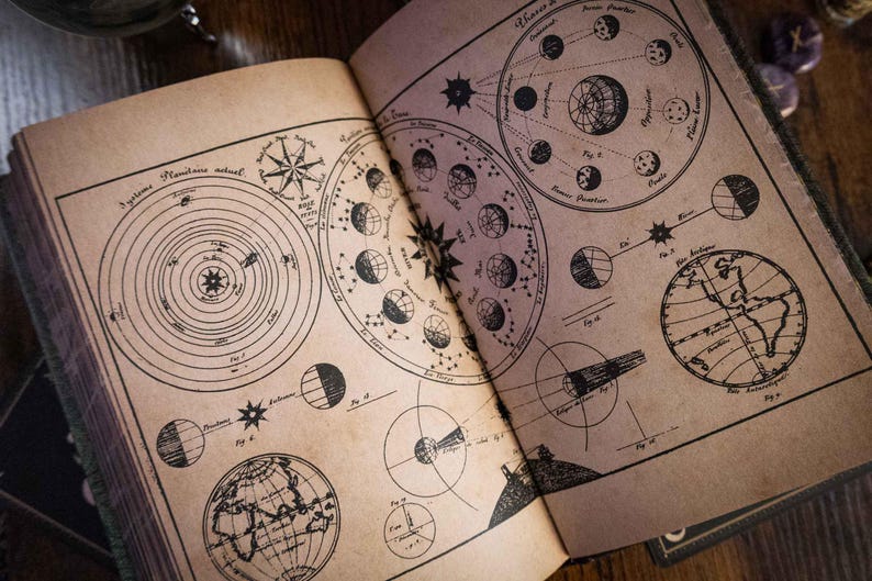 K&ouml;nnte beinhalten: Ein aufgeschlagenes Buch mit gealterten, beigefarbenen Seiten, das schwarze Tuschezeichnungen astronomischer Diagramme zeigt, darunter Planetenbahnen, Mondphasen und Globen. Der Text enth&auml;lt Beschriftungen wie 'Syst&egrave;me Plan&eacute;taire actuel' und verschiedene Abbildungsnummern.
