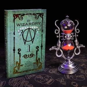May include: A green hardback book titled The Wizardry Omnibus Volume 1 with gold lettering and a metallic silver hourglass featuring dragon-shaped side supports and bright orange sand inside the glass bulbs, standing on a circular base.