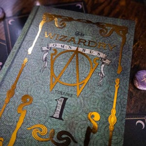 K&ouml;nnte beinhalten: Ein gebundenes Buch mit dem Titel The Wizardry Omnibus Volume 1 mit einem strukturierten blaugr&uuml;nen Einband, der goldene Folienakzente, ein zentrales Emblem, dekorative stabartige Motive und den Titel in goldener Schrift aufweist.