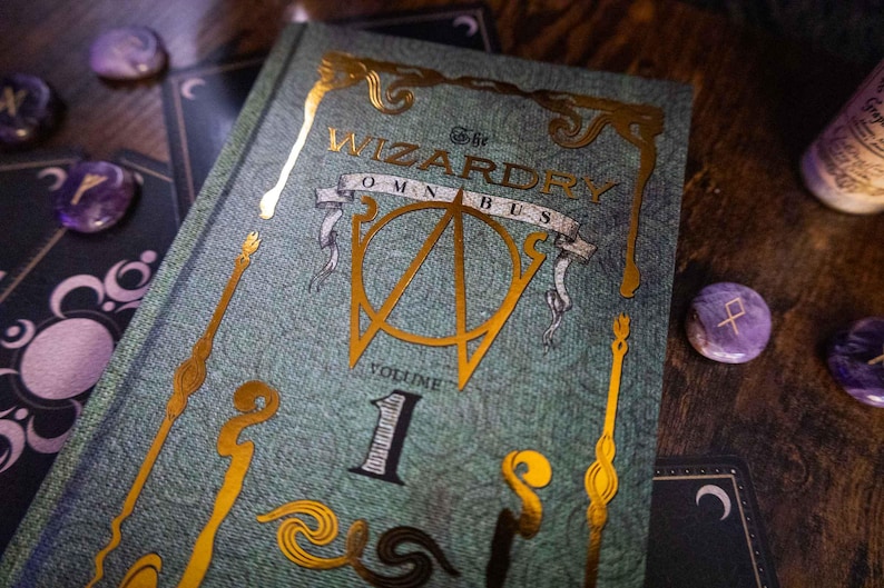 K&ouml;nnte beinhalten: Ein gebundenes Buch mit dem Titel The Wizardry Omnibus Volume 1 mit einem strukturierten blaugr&uuml;nen Einband, der goldene Folienakzente, ein zentrales Emblem, dekorative stabartige Motive und den Titel in goldener Schrift aufweist.