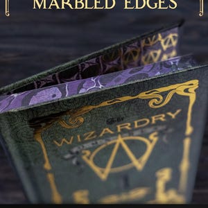 K&ouml;nnte beinhalten: Ein dunkelgr&uuml;nes Hardcover-Buch mit goldener Schrift, die WIZARDRY liest, und einem zentralen goldenen Emblem. Das Buch hat violett und schwarz marmorierte Buchschnitte. Der Text DELUXE MARBLED EDGES und ARCHIVEOFWIZARDRY.COM ist zu sehen.