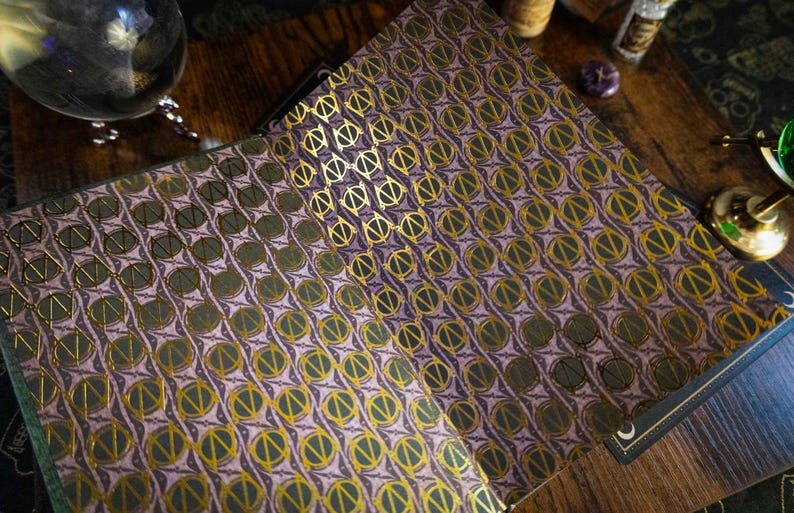 K&ouml;nnte beinhalten: Ein aufgeschlagenes Buch mit Vorsatzpapier, das ein sich wiederholendes Muster aus goldenen kreisf&ouml;rmigen Symbolen auf einem ged&auml;mpften violetten Hintergrund zeigt. Das Design besteht aus einer vertikalen Linie, die einen Kreis teilt.
