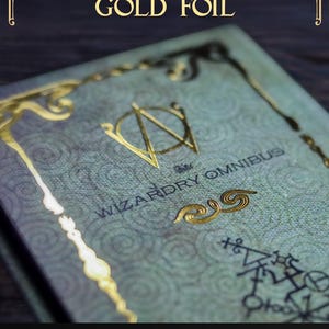 K&ouml;nnte beinhalten: Ein gr&uuml;ner Bucheinband mit dem Titel WIZARDRY OMNIBUS und Goldfolienverzierungen, darunter ein zentrales Logo, dekorative R&auml;nder und geometrische Symbole. Der Text EMBELLISHED with GOLD FOIL und ARCHIVEOFWIZARDRY.COM sind auf dem Einband zu sehen.