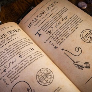 K&ouml;nnte beinhalten: Ein aufgeschlagenes Buch mit gealterten, beigefarbenen Seiten, die mit schwarzem Text und Illustrationen bedruckt sind. Die linke Seite ist mit Spirit Guard Charm und die rechte mit Disarming Charm betitelt. Beide Seiten enthalten detaillierte Beschreibungen, magische Symbole und Diagramme.