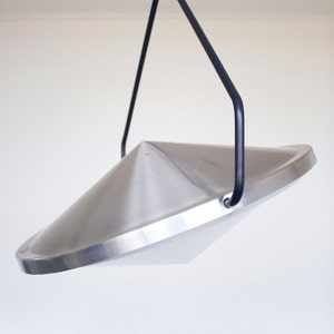Puede incluir: Una lámpara colgante de metal plateado con una pantalla en forma de cono y una varilla de suspensión de metal negro.
