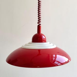 Może przedstawiać: Czerwona lampa wisząca z białym pierścieniem akcentującym i spiralnym przewodem. Oprawa oświetleniowa ma błyszczące wykończenie i zaokrąglony klosz. Przewód ma pasujący czerwony kolor i jest zwinięty. Światło jest zawieszone pod sufitem.