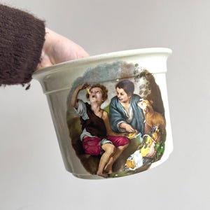 May include: Cream-colored ceramic planter with a decorative illustration of two people and a dog. The illustration features figures in period clothing, with a warm color palette. The planter has a slightly flared rim and a cylindrical shape.