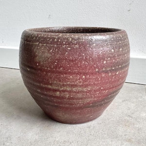 Könnte beinhalten: Ein runder Keramik-Pflanztopf mit einer weiten Öffnung. Die Außenseite hat eine gesprenkelte, rotbraune Glasur mit Bändern aus hellem Braun und Beige. Der Pflanztopf steht auf einer hellgrauen Fliesenoberfläche, und der Hintergrund ist eine weiße Wand.