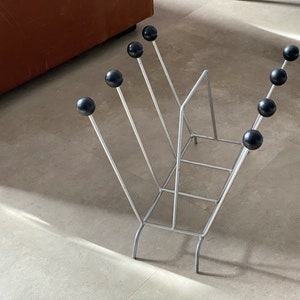 May include: A silver metal magazine rack with black round knobs on the ends of each arm. The rack is standing on a light gray tiled floor.