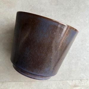 Könnte beinhalten: Eine braune Keramiktasse mit blauer Glasur. Die Tasse ist leicht geneigt und die Glasur hat einen marmorierten Effekt.